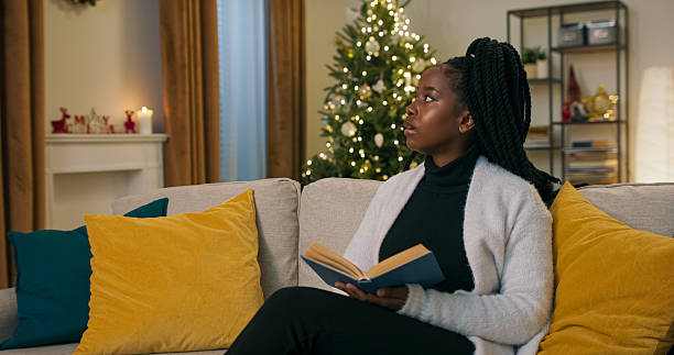 istock A young woman sits on a sofa, reading a book with a blue cover. After reading a section, she stops to reflect, looking dreamily out the window. Pillows rest on the sofa nearby. 2239538030