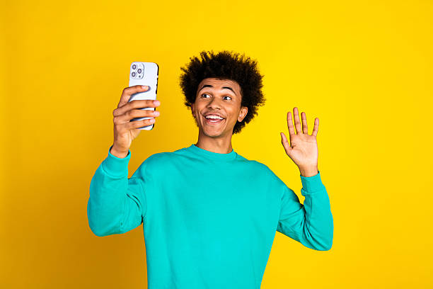 istock Young man with curly hair smiling and gesturing while taking a selfie on a smartphone against a vibrant yellow background 2239543749