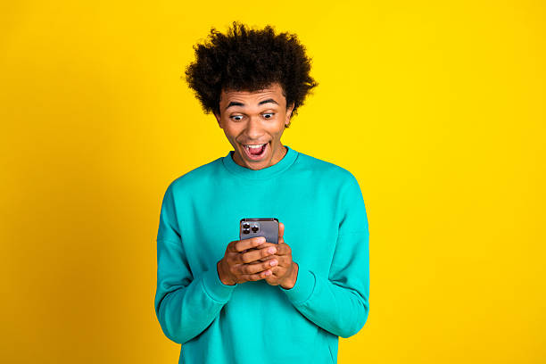 istock Cheerful young man in teal sweatshirt reacts energetically while using smartphone against vibrant yellow background 2239543750