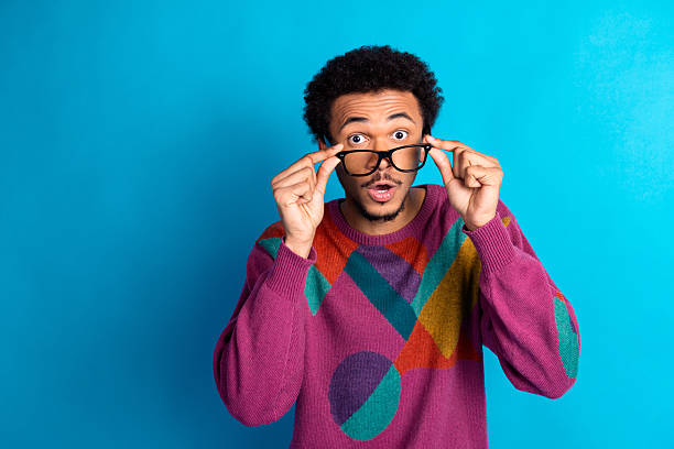 istock Young man in colorful retro sweater with surprised expression adjusting glasses against a vibrant blue background 2239549098