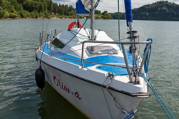 Czorsztyn Lake, Poland – August 6, 2025: A compact white sailboat with blue deck design is moored near the shore. Calm lake water reflects sunlight as forested hills rise in the background.
