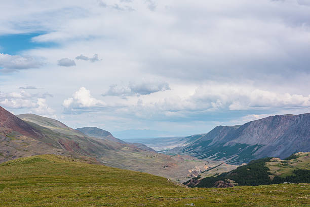 istock Dramatic top view from green pass through sunlit forest hills to wide alpine valley between large mountain range and multicolor mountains under cloudy gray sky. Big rocky ridge in changeable weather. 2239577426