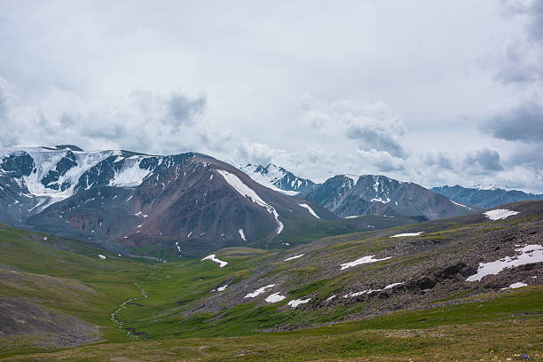 istock Dramatic view to green alpine valley with long creek and large snow-capped mountain range under cloudy gray sky. Mountain stream among rock hills with snowfields and high mountains. Awesome landscape. 2239580284