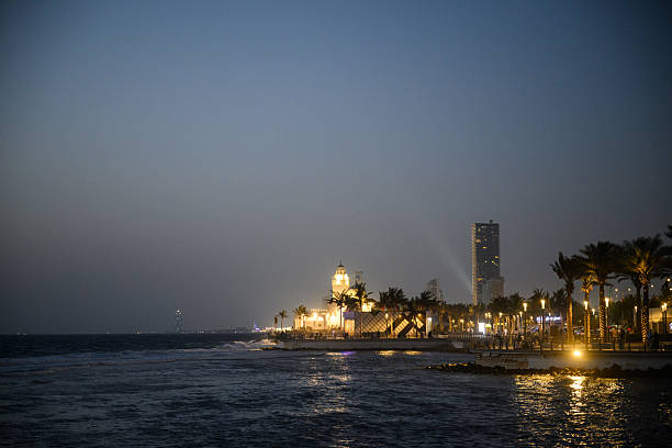 Night time coastal view of the Jeddah Corniche showing glowing street lights, modern towers and reflections sparkling across the water under a blue sky.