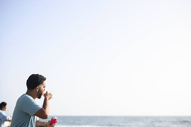 Side view of a young man enjoying ice cream while looking out over the sea, framed against a clear blue sky and calm water on a coastal promenade.