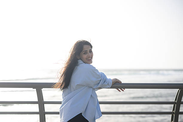 Portrait of a young woman smiling at camera while leaning casually against a metal railing, with sunny ocean waves and open sky creating a peaceful coastal backdrop.