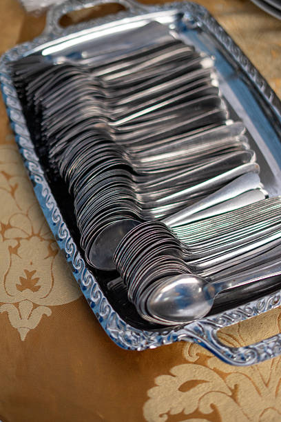 A large quantity of clean, shiny stainless steel spoons neatly stacked on a decorative silver-colored tray, ready for a catering event or formal dining service.