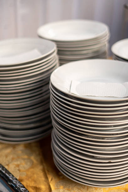Multiple stacks of clean, white ceramic plates with dark rims are neatly arranged on a patterned golden tablecloth, ready for use at a catering event, buffet, or restaurant service.