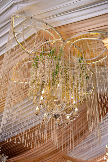 Elegant chandelier and festive ceiling decor with drapes, hanging artificial flowers, and sparkling string lights, perfect for weddings and celebratory events.