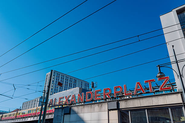 A bold red Alexanderplatz sign dominates the foreground as tram lines cross a clear blue sky. Modern buildings frame the scene, capturing urban life, transit energy, and Berlin’s iconic cityscape.