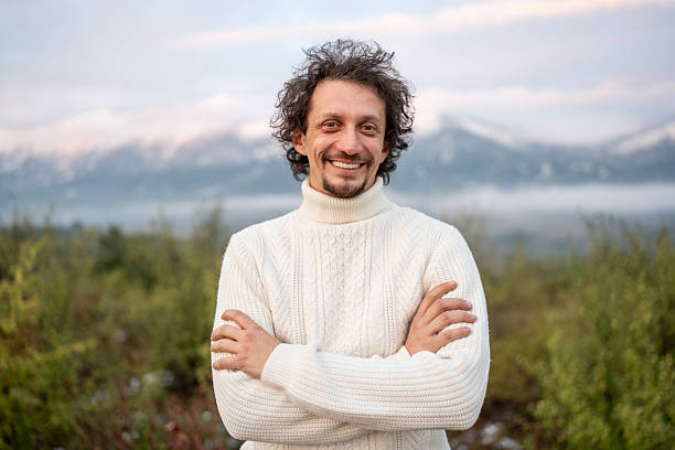 A cheerful man stands outdoors in a white cable-knit sweater, arms crossed and smiling, with a scenic mountain range and soft sky in the background. Ideal for lifestyle, outdoor, and fashion stock uses.