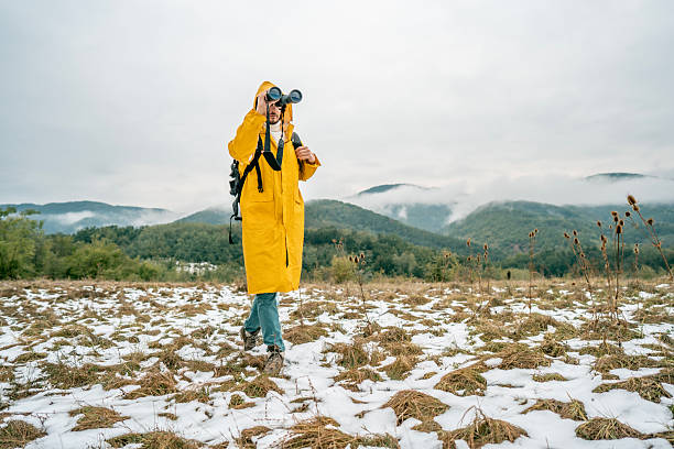 A lone explorer in a bright yellow raincoat and backpack stands in a snowy meadow, peering through binoculars toward distant mountains. An outdoor adventure scene filled with curiosity and resilience.