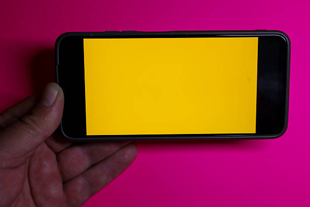 ​A hand holds a smartphone in horizontal orientation with a bright yellow screen against a saturated pink background. This dynamic shot with strong color contrast is ideal for a mockup showcasing a website, graphics, or applications related to fashion, technology, and social media