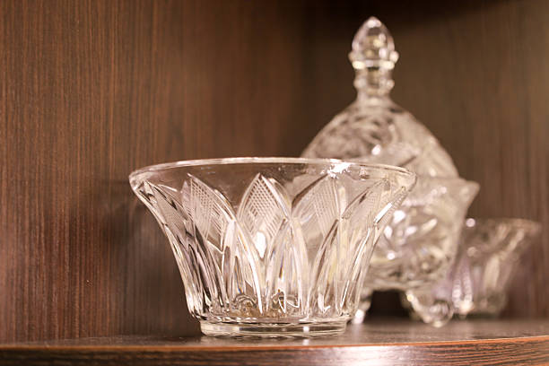 istock ​A clear crystal vase with an embossed pattern stands on a wooden shelf, displayed in a brown cabinet along with other glass items. Light reflects off the facets, emphasizing the decorative and elegant design of the tableware. 2239592288