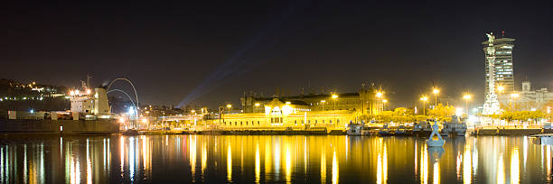 Panoramic view of Barcelona's Port Vell at night, featuring bright reflections of city lights on the water and the iconic Columbus Monument framing the city skyline