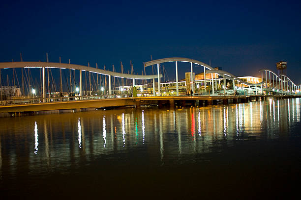 Rambla de Mar bridge illuminated by city lights creating vibrant reflections on the calm water of Port Vell marina, showing nautical masts under a dark twilight sky in Barcelona, Spain