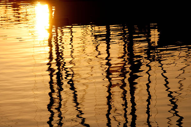 Warm golden sunset light shimmering on rippled harbor water in Barcelona, creating abstract patterns and dark vertical reflections of boats and masts for a serene, textured scene