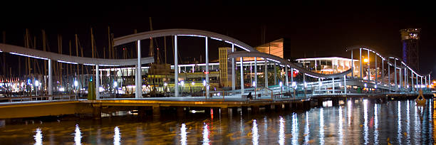 Rambla de Mar pedestrian bridge extending over the water in Barcelona's Port Vell at night, featuring modern architecture and urban reflections from city lights