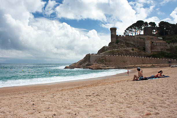 Tosa de Mar, Cataluña, Spain - April 23, 2011: People enjoying a relaxing day on the sand beach by the Mediterranean sea with the historic Vila Vella castle and defensive walls in Tossa de Mar, Catalonia, Spain, on a sunny day
