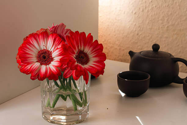 Arrangement of red and white gerbera daisies in clear glass jar with water, placed beside dark teapot and cups on white surface. Warm lighting creates cozy atmosphere, perfect for decorative, lifestyle, and editorial projects.