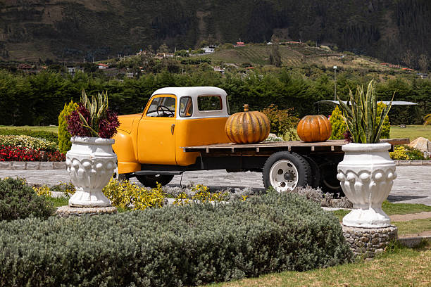 An antique farm truck carrying large pumpkins on its platform, resting outdoors on a stone patio with hills rising in the distance.
