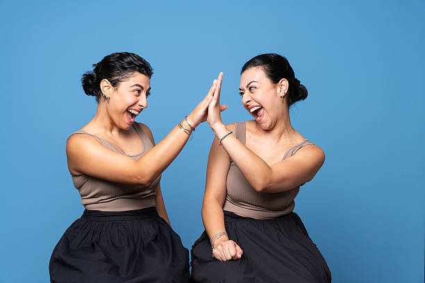 Studio portrait of two smiling women in matching outfits sitting and celebrating with a cheerful high five gesture against blue background.