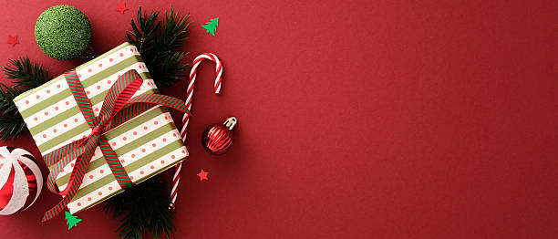 istock Christmas gift box with striped ribbon surrounded by ornaments, candy canes, pine branches, and confetti on red background. Festive Xmas banner design. 2239614085