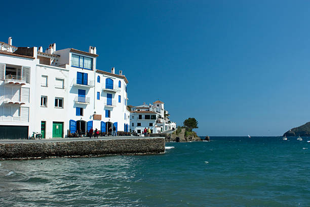 Village of Cadaques in Catalonia, Spain, showcasing traditional whitewashed buildings with vibrant blue shutters lining the waterfront promenade under a clear sky