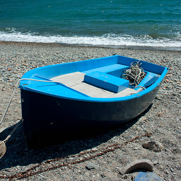 Small blue fishing boat pulled ashore on the pebble beach of Cadaques, Catalonia, with turquoise Mediterranean Sea waves gently breaking in the background on a sunny day