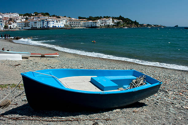 Blue small boat resting on the pebble beach of Cadaques with the white village and turquoise Mediterranean sea in the background, a popular tourist destination