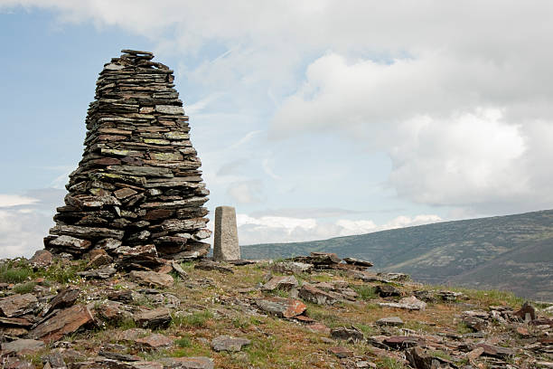 Stone cairn and a surveying marker standing on a rocky mountain summit, with green hills and a cloudy sky in the background, capturing a remote and peaceful rural landscape