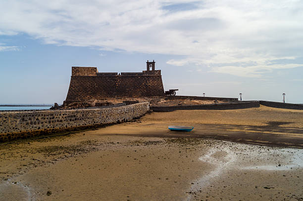 A panoramic view of Castillo de San Gabriel, a well-preserved 16th-century stone fortress in Arrecife, Lanzarote. The sandy low tide foreground with a beached blue-white rowboat and dramatic sky conveys the historical and coastal atmosphere of the Canary Islands.