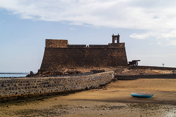 The historic Castillo de San Gabriel fortress stands watch over the sandy shore and a blue rowboat during low tide. The volcanic stone walls, clear sky, and canons evoke memories of Lanzarote’s strategic maritime past.