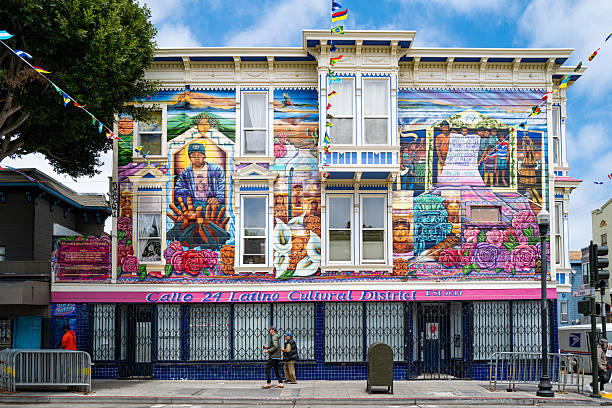 San Francisco, CA - U.S.A. - May 24, 2025: Two people walk down 24th Street in front of a colorful building with mural by lead muralist Daniel Galvez that is part of the Calle 24 Latino Cultural District area in the Mission District.