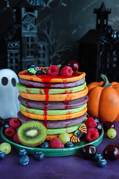 Image of Halloween food, multicoloured pancake stack served with fresh fruit, alternating orange, purple, green layers of flapjacks, raspberry, blueberry, dripping strawberry sauce, spiders, ghost, pumpkin, spooky house background, focus on foreground