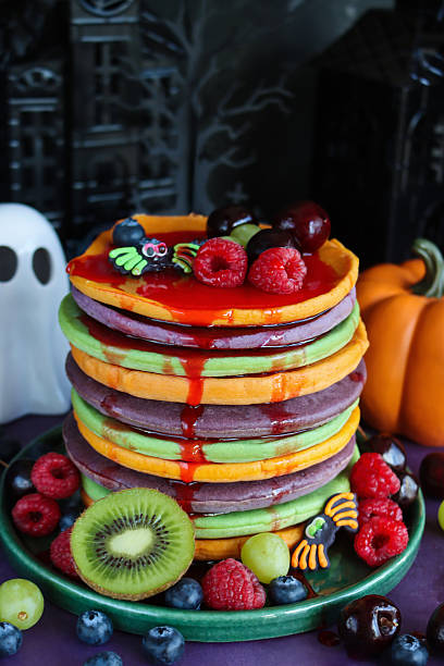 istock Close-up image of Halloween food, multicoloured pancake stack with fresh fruit, alternating orange, purple, green layers of flapjacks, raspberry, blueberry, dripping strawberry sauce, fondant spiders, china ghost, pumpkin, spooky house background 2239647839