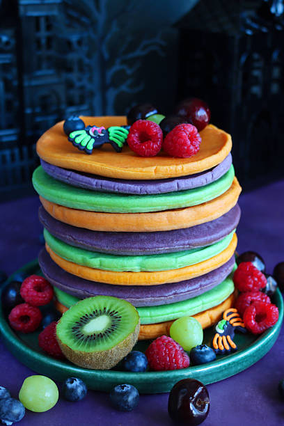 istock Close-up image Halloween food, stack of multicoloured pancakes served with fresh fruit, alternating orange, purple, green layers of flapjacks, raspberry, blueberry, kiwi, grapes, cherries, fondant spiders, spooky house background, focus on foreground 2239648042