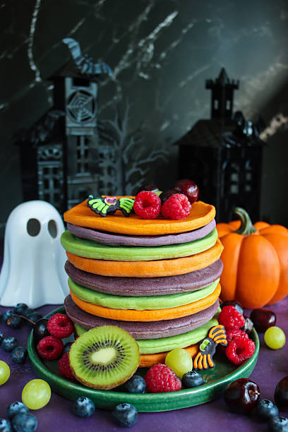 istock Image of Halloween food, multicoloured pancake stack with fresh fruit, alternating orange, purple, green layers of flapjacks, raspberries, blueberries, fondant icing spiders, pumpkin, white china ghost, spooky house  background, focus on foreground 2239648064