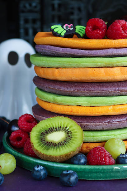 istock Full frame image of Halloween food, multicoloured pancake stack with fresh fruit, alternating orange, purple, green layers of flapjacks, raspberries, blueberries, fondant icing spider, white china ghost, spooky house  background, focus on foreground 2239648139