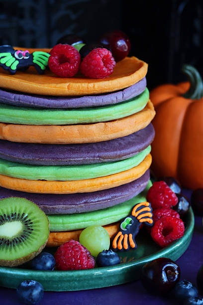 istock Full frame image Halloween food, stack of multicoloured pancakes served with fresh fruit, alternating orange, purple, green layers of flapjacks, raspberry, blueberry, fondant icing spiders, pumpkin, dark background, focus on foreground 2239648150