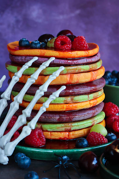 istock Image of Halloween food, skeletal hand reaching for multicoloured pancake stack and fresh fruit, alternating orange, purple, green layers, raspberries, blueberries, cherries, dripping strawberry sauce, mottled purple background, focus on foreground 2239648402