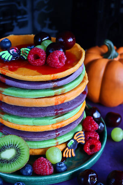 istock Close-up image of Halloween food, multicoloured pancake stack, fresh fruit, alternating orange, purple, green layers of flapjacks, raspberry, blueberry, dripping strawberry sauce, fondant icing spiders, pumpkin, dark background, focus on foreground 2239649325