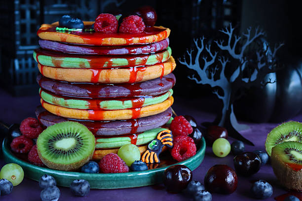 istock Close-up image Halloween food, multicoloured pancake stack with fresh fruit, alternating orange, purple, green layers of flapjacks, raspberry, blueberry, dripping strawberry sauce, fondant icing spiders, 3D paper craft tree, spooky house background 2239649591