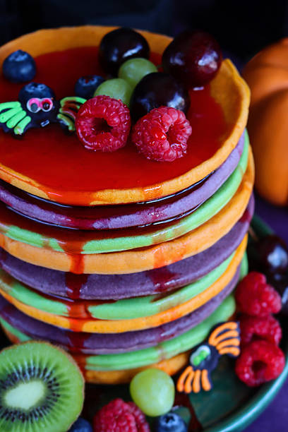 istock Full frame image Halloween food, stack multicoloured pancakes, fresh fruit, alternating orange, purple, green layers of flapjacks, raspberry, blueberry, dripping strawberry sauce, fondant icing spiders, dark background, elevated view, focus on foreground 2239649592
