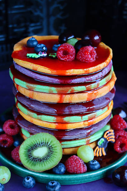 istock Close-up image Halloween food, stack of multicoloured pancakes served with fresh fruit, alternating orange, purple, green layers of flapjacks, raspberry, blueberry, dripping strawberry sauce, fondant icing spiders, dark background, focus on foreground 2239649761