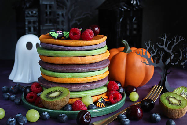 istock Image Halloween food, stack of multicoloured pancakes served with fresh fruit, alternating orange, purple, green layers of flapjacks, raspberry, blueberry, fondant icing spiders, ghost, pumpkin, 3D paper craft tree, spooky house, focus on foreground 2239649892