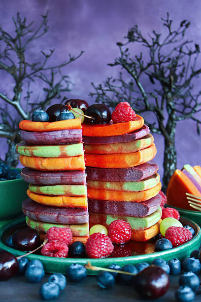 istock Image of Halloween food, sliced multicoloured pancake stack and fresh fruit, alternating orange, purple, green layers of flapjacks, raspberries, blueberries, cherries, dripping strawberry sauce, model trees, mottled purple background, focus on foreground 2239649946
