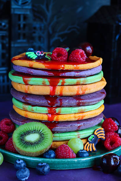 istock Close-up image Halloween food, stack of multicoloured pancakes served with fresh fruit, alternating orange, purple, green layers of flapjacks, raspberry, blueberry, dripping strawberry sauce, fondant icing spider, dark background, focus on foreground 2239650102
