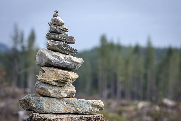 a small stack of volcanic rocks, known as a cairn, carefully balanced on top of each other along a hiking trail in Kali Talang. The stones vary in size and shape, with rough, porous textures and patches of moss clinging to their surfaces, adding a natural, weathered charm. In the background, more stacked rocks can be seen, blending harmoniously with the lush green foliage of the surrounding forest. These cairns not only serve as trail markers for hikers but also reflect the tranquil and meditative atmosphere of the natural landscape.