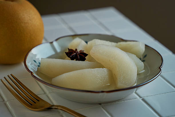 A calm, home-style dessert of Japanese Asian pears (nashi) gently poached in light sugar syrup infused with star anise, served chilled in a small ceramic bowl. Natural light and minimal styling make it ideal for autumn menus, recipes, and seasonal features.